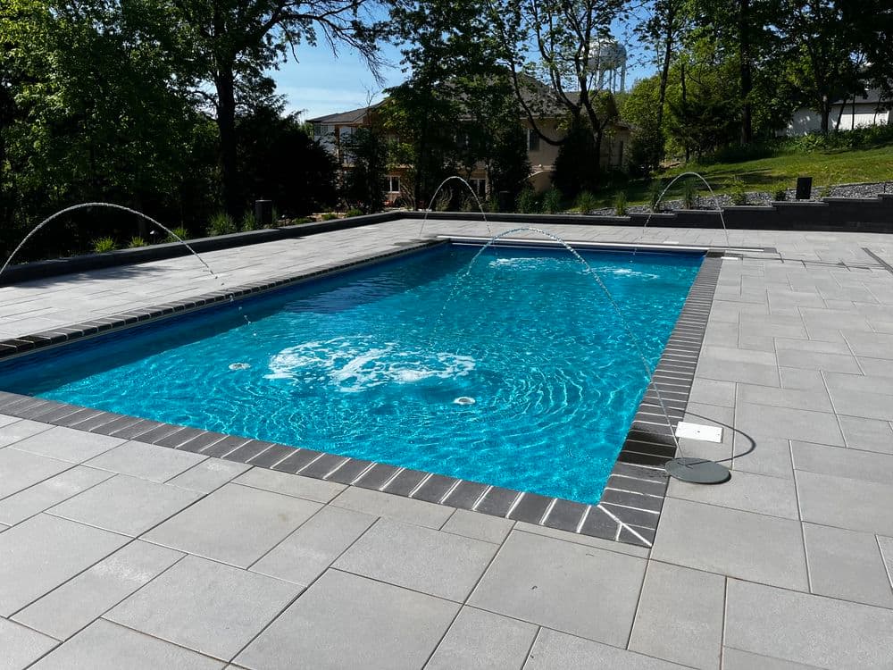 Modern rectangular swimming pool with water features and gray stone patio surrounded by trees.