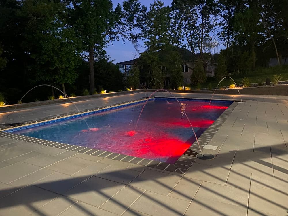 Illuminated backyard swimming pool at night with colorful fountain features and lush greenery.