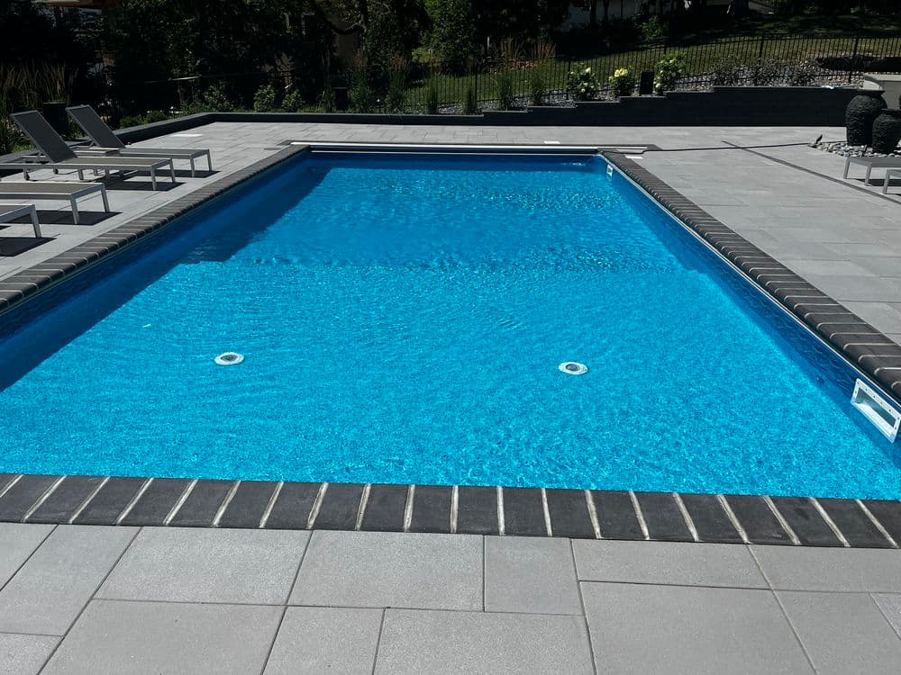 Crystal clear swimming pool with gray tile surround and lounge chairs in a landscaped yard.