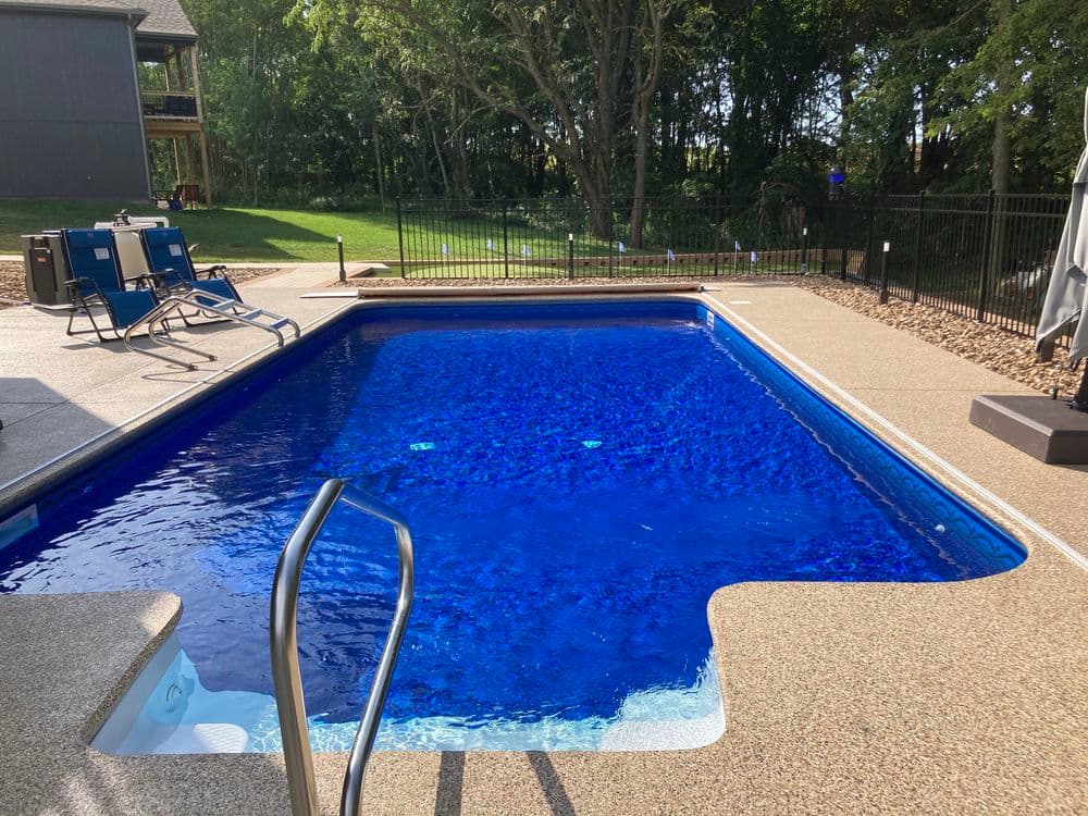 Inground swimming pool with clear blue water, surrounded by a patio and green trees.
