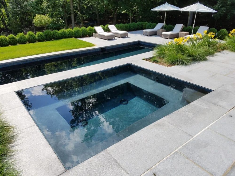 Modern backyard oasis with a spa, pool, and lounge chairs surrounded by lush greenery.